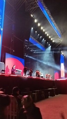 The video captures a live musical performance on a large, brightly lit stage at night, likely in Maipú, Chile. The stage features multiple levels and is equipped with extensive lighting rigs, powerful spotlights, and smoke/haze effects, creating a dynamic atmosphere. Blue and red decorative elements adorn the stage structure.

Several performers, some wearing red tops, are visible on stage, appearing to be musicians or singers. In the foreground, camera operators are seen recording the event. The audio features energetic music with singing in Spanish, including the phrases "¡Catracho, catracho!" and "usted me promete." The overall scene suggests an engaging concert or live event with a vibrant visual and auditory experience. No visible text is discernible in the video.