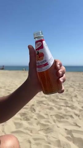 A person on a sunny beach holds up a bottle of orange basil seed drink and takes a sip. The bottle has a red and white label with an image of an orange on it. The beach has golden sand, with the blue ocean and sky in the background. A few people are scattered on the beach and in the water. There's also a white beach umbrella visible in the background. The person holding the drink is wearing a black shirt.