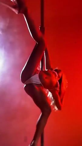 A woman in a sparkling bikini performs a pole dance in a dimly lit room with red and purple lighting and smoke effects. She is shown in various poses, including hanging upside down, bending her body, and climbing the pole.
