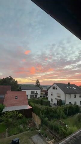 The video shows a panoramic view of a suburban neighborhood during sunset. The sky is filled with dramatic clouds that are illuminated by the setting sun, creating a vibrant display of orange, pink, and yellow hues. Several houses with tiled roofs and green gardens are visible in the foreground and middle ground. The overall atmosphere is peaceful and serene, capturing the beauty of a natural phenomenon in a residential setting. There are no people or animals visible in the video, and the focus is entirely on the landscape and the sky. The camera pans slowly across the scene, offering a sweeping view of the sunset.