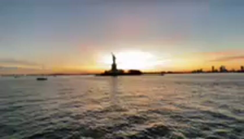 This media file captures a serene view of the Statue of Liberty at sunset. The iconic monument stands silhouetted against a vibrant sky painted with hues of orange, pink, and yellow, indicative of the twilight hours. The sun, a bright orb, is positioned behind the statue, creating a dramatic halo effect.

The scene is set on a body of water, likely New York Harbor, with gentle ripples suggesting a calm atmosphere. In the distance, the New York City skyline is faintly visible on the right, adding depth to the panorama. A few sailboats are scattered across the water, further enhancing the peaceful ambiance.

The overall mood is one of tranquility and awe, as the natural beauty of the sunset complements the grandeur of the Statue of Liberty. The time of day is clearly sunset, characterized by the warm, low-angled light and the colorful sky. The weather appears to be clear, allowing for an unobstructed view of the sunset and the cityscape.

There is no visible text within the media file.