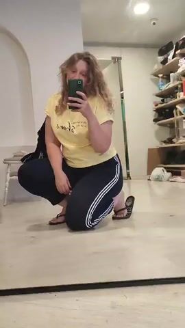 This is a video of a person taking a selfie in a room. The person is wearing a yellow t-shirt and navy blue pants with white stripes. They are in a squatting position, holding a smartphone. The room appears to be a studio or storage area, with photography equipment such as softboxes, tripods, and a computer visible in the background. There are also shelves with items on them and a white archway. The floor is light-colored wood.