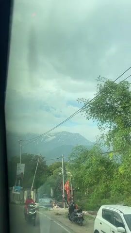 The view from inside a vehicle moving along a road captures a scenic landscape. The sky is overcast with a mix of clouds and patches of blue. In the distance, snow-capped mountains stand out against the sky, partially obscured by the clouds. Below the mountains, there are green trees and vegetation. Utility poles with wires crossing the sky are visible, partially obscuring the mountain view. On the side of the road, there's a sign with a portrait of a man advertising 'LASSI'. A woman in a pink jacket is riding a scooter on the road. Another parked scooter is visible near a small structure and flags on the roadside.