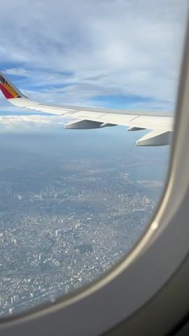 This is a vertical, first-person perspective video taken from inside an airplane, looking out the window. The airplane's wing is visible in the upper left portion of the frame, featuring a red, yellow, and blue logo at the wingtip. Below the wing, a vast urban landscape stretches out, densely packed with buildings of varying heights, suggesting a large city. The cityscape appears somewhat hazy due to atmospheric conditions. In the distance, a body of water, likely a bay or ocean, can be seen bordering the city. The sky above is a mix of blue and white clouds, with sunlight illuminating the scene. The video captures the smooth, steady movement of the airplane as it flies over the city. There are no people or interactions visible, only the view from the aircraft.