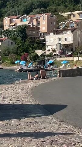 The video shows a scenic view of a coastal town, likely Donja Lastva, Montenegro. In the foreground, a stone-paved walkway curves away from the camera, meeting an asphalt road. The walkway overlooks a body of water, likely the Adriatic Sea, where several people are visible. Two individuals, a man and a woman, are seated on the edge of the walkway, facing the water. The man is seated with his back to the camera, while the woman is lying down beside him.

Further back, along the waterfront, are several blue umbrellas providing shade for beach chairs. Other individuals are seen relaxing on the shore or wading in the water. Behind the waterfront, a cluster of multi-story buildings, painted in various pastel colors, are nestled on a lush green hillside. These buildings appear to be residential homes, with balconies and windows visible. The overall setting suggests a sunny day, characteristic of a summer vacation spot. The atmosphere appears relaxed and leisurely, with people enjoying the coastal environment. No visible text is present in the video.