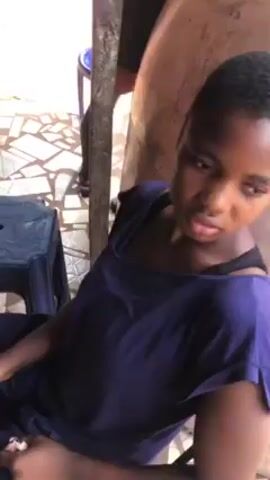 A young, dark-skinned woman is seated in a chair, wearing a dark blue short-sleeved shirt and a black bra strap is visible. She has short, cropped dark hair. The woman is looking down with a slight frown on her face. Behind her is a large, light-colored barrel-shaped object. There is a grey wooden post on the left side of the frame and a blue plastic stool is partially visible next to the post. The ground appears to be tiled with geometric patterns. Another person is partially visible standing behind the barrel.