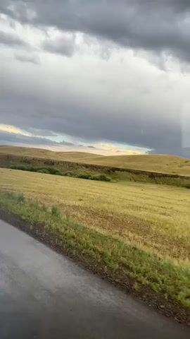 The video shows a view from a vehicle moving along a wet asphalt road. To the right of the road is a field of dry, golden grass with some green patches. Further into the distance, there is a low, rolling hill covered in grass. Above the hill, a dark, cloudy sky looms, with lighter clouds and a hint of blue sky visible near the horizon. The overall impression is of a somewhat overcast or approaching storm.