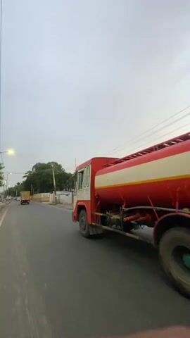This video captures a street view in St. Thomas Mount-Pallavaram Cantonment Board, India. The primary subject is a red and white water tanker truck moving along a paved road. The truck has a distinct design with colorful decals on its cab and a large cylindrical tank. Yellow and red accents are visible on the truck's chassis and piping.

In the background, a white gate and a building with a sign that reads "Shivoham" are visible on the left side of the road, suggesting a commercial or industrial establishment. Lush green trees line the street, indicating a somewhat suburban or semi-rural environment. The sky is overcast, suggesting a cloudy or possibly impending rain. The lighting indicates it is either early morning or late afternoon.

The camera appears to be mounted on another moving vehicle, capturing the truck from a side-angle as it drives. The overall impression is a glimpse of daily life and transportation on a road in this Indian cantonment board. No people are clearly visible in the frame, and the focus is entirely on the truck and its surroundings. The presence of utility poles and overhead wires further emphasizes an urban or semi-urban setting.