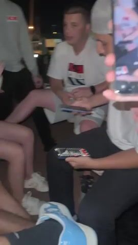 This video shows a group of young adults sitting together outdoors at night. The individuals are casually dressed in t-shirts and shorts. One person is holding a phone and appears to be showing something on it to another person who is holding a remote control. The atmosphere seems relaxed and friendly. The background is dimly lit, suggesting it is evening or night.
