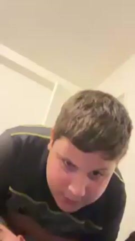 A young boy wearing a patterned gray and black short-sleeved shirt looks down at the camera, sticks out his tongue, then smiles. He stands up and turns to his right, slightly out of view, then turns back to face the camera, smiling.  The background consists of a white wall and part of a white door frame. Someone is lying down below the boy in the frame, but only the top of their head is visible.