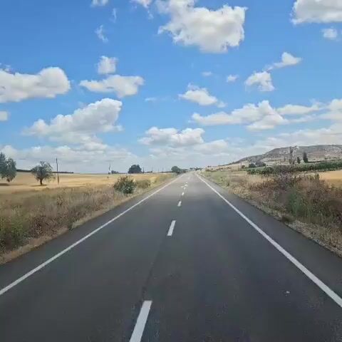 The video shows a drone shot of a highway in a rural area. The highway is asphalt with white dashed lines in the center and solid white lines on the sides. The road is flanked by dry, yellowish grass and some green bushes. In the background, there is a hill with some vegetation and a few scattered buildings. The sky is blue with scattered white clouds. The camera pans slightly to the right, revealing more of the landscape and the hill. A large white arrow shape is visible on the side of the hill.
