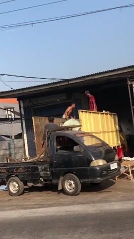 The video shows three men working on a black flatbed truck parked in front of a building with a corrugated metal roof and blue metal doors. The truck is loaded with various items, including what appears to be scrap metal, wood, and possibly furniture. One man, wearing a red shirt, is standing on the truck bed and appears to be directing the loading. Another man, shirtless, is also on the truck bed, handling some of the materials. A third man, wearing a dark shirt, is standing on the ground next to the truck, helping to load items. The environment appears to be an outdoor work area, possibly a junkyard or a recycling facility, with debris and other discarded items visible on the ground. The sky is clear and blue, suggesting daylight. The scene is captured from a slightly elevated angle, looking across a paved surface.