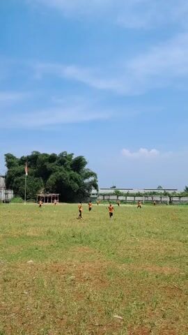 A soccer game is being played on a grassy field under a blue sky with scattered clouds. Several players wearing orange vests are scattered across the field. A large bamboo clump is in the background, with a flag of Indonesia next to it. A small boy wearing a striped shirt is watching the game.