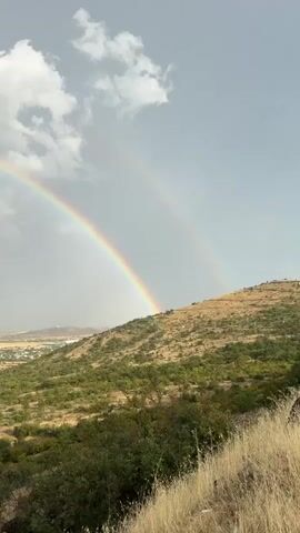 The video shows a panoramic view of a landscape with a double rainbow stretching across a cloudy sky. In the foreground, there is a grassy hill with scattered green bushes. Beyond the hill, a town or city is visible, with buildings and roads. The sky is a mix of grey clouds and patches of blue, with the sun faintly visible behind the clouds. The double rainbow is vibrant, with distinct bands of color. The video pans slightly to the right, revealing more of the town and the surrounding terrain. The overall impression is one of natural beauty and a recent or ongoing rain shower.
