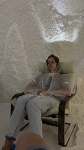 A group of people are sitting in comfortable chairs within a salt room. The walls and ceiling are covered in white salt crystals, giving the impression of a cave. The lighting is subdued. Most individuals are wearing light-colored robes and blue shoe covers. Some are holding their phones, while others appear to be resting with their eyes closed. One person in the foreground is partially visible, facing away from the camera. The atmosphere seems calm and relaxing.