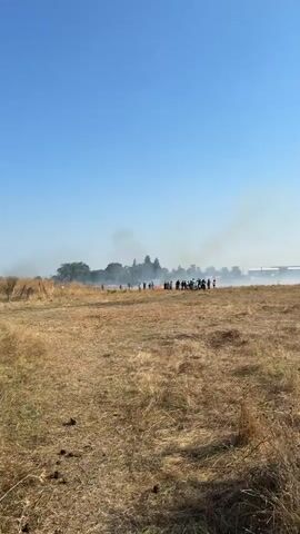 A group of people are gathered in a dry, grassy field under a clear blue sky. Smoke is visible in the distance, rising from a fire in the field. Some of the people are observing the fire, while others appear to be engaged in some activity around it. There are also bicycles present among the group. The landscape is dry and arid, suggesting a hot and dry environment. The overall scene depicts a community event or gathering centered around a fire in an open field.