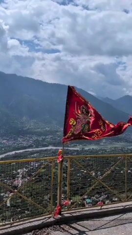 A red flag with an image of a Hindu goddess and text in Hindi is affixed to a pole in the foreground. The flag is partially obscured by the pole and is billowing in the wind. Behind the flag, a valley with a river and a town is visible, surrounded by green mountains. The sky is cloudy with patches of blue. The flag appears to be made of fabric and is attached to the pole with some red ribbons. There is a yellow railing in the foreground.