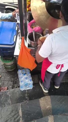 A man wearing a white shirt and a red apron is preparing food at an outdoor stall. He is using a large metal pan and appears to be assembling a sandwich or wrap. Several bottles of water are stacked on the ground near him, along with a blue cooler and some buckets. The stall has various ingredients and containers visible, and the overall setting appears to be a street or market.