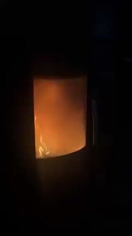 The video shows a close-up of a wood-burning stove with a fire visible inside. The stove is dark, and the only light source is the orange glow from the flames and burning embers within the stove. There is no visible movement of the camera. There is the sound of a dog panting.