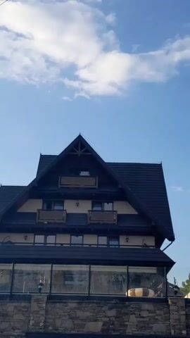 The video shows the upper facade of a dark-roofed building with multiple stories and balconies against a bright blue sky with scattered white clouds. The architecture appears to be traditional or rustic, with wooden accents and a steeply pitched roof. A stone wall and a glass-enclosed area are visible at the bottom of the frame. There are no people or active events captured in the footage.