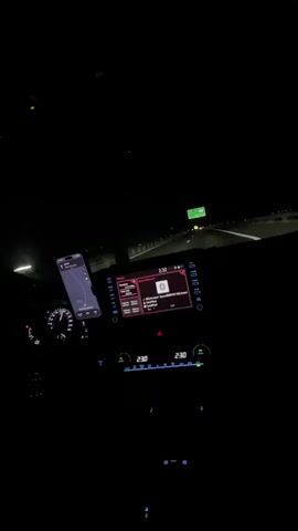 The video shows the interior of a car at night, traveling on a highway. The dashboard is visible, with the infotainment system displaying music information and navigation. The driver's phone, mounted on the windshield, also shows navigation. The headlights illuminate the road ahead, and other cars can be seen in the distance. The climate control settings are visible on the dashboard, showing 23 degrees Celsius. The overall impression is one of a nighttime drive.