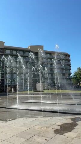 A static, vertical video captures a contemporary fountain with multiple jets of water spraying upwards. The fountain is set in a paved urban area with square-shaped stone tiles. Behind the fountain, a large, multi-story building with a curved facade and numerous balconies stands under a clear blue sky. A statue is visible behind the water jets, in front of the building. The sunlight casts shadows on the paving stones. The video appears to be filmed on a bright, sunny day.