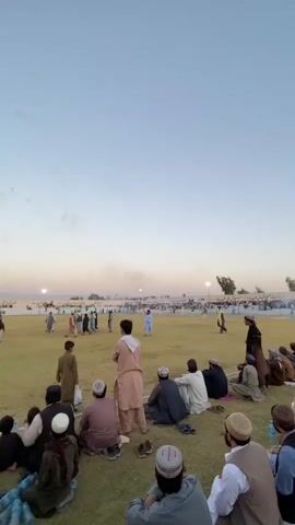 The video captures an outdoor event, possibly a sporting match or festival, at dusk. A crowd of spectators is seated on a grassy field, facing a distant playing area. The sky is a gradient of pale blue and soft orange, indicative of sunset. In the foreground, people of various ages are dressed in traditional attire, with many wearing skullcaps. They are sitting in small groups, some conversing, others watching the event unfold. On the field, numerous individuals are visible, some actively participating in an activity, while others stand and observe. The background reveals a long line of tents or structures, and further in the distance, a faint mountain range. Several lights are illuminated around the playing area, suggesting the event continues into the evening. The overall atmosphere appears relaxed and communal.