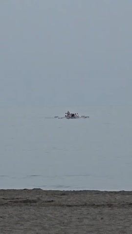The video shows a wide shot of a calm ocean under an overcast sky. In the middle ground, a small boat with several people is visible. The boat appears to be a rowing boat with multiple oarsmen. In the foreground, a sandy beach is visible. A yellow buoy is floating in the water to the right of the boat. The boat is moving from left to right across the frame.