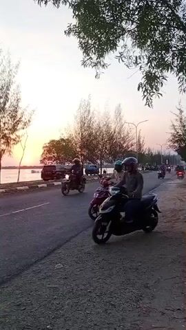The video shows a group of people riding motorcycles along a paved road next to a body of water. The sun is setting, casting a warm glow over the scene and silhouetting trees along the roadside. The motorcycles are varied in style, with some appearing to be scooters and others more traditional motorcycle models. Several individuals are visible on the motorcycles, some riding alone and others with passengers. The road appears to be a public thoroughfare, with other vehicles and possibly buildings visible in the distance. The overall atmosphere is one of leisurely activity and enjoyment of the evening scenery.