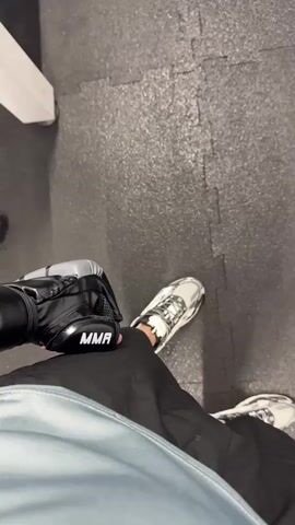 A person stands on a grey, interlocking tile floor, holding a black and grey MMA glove in their hand. They are wearing black pants and white shoes with black accents. The camera pans up to show a black RDX punching bag hanging from a chain against a white wall.