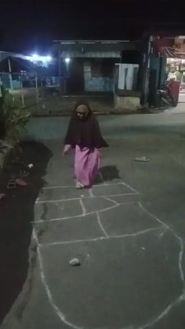A young girl in a hijab and pink skirt is playing hopscotch at night. The hopscotch court is drawn on an asphalt surface with chalk. The girl throws a small stone into one of the squares, then hops through the court. There are some buildings and streetlights in the background, indicating an outdoor urban or suburban environment.