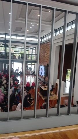 This is a horizontal, slightly angled, high-angle shot looking through horizontal metal bars at a large gathering of people, possibly in a mosque or community hall. The majority of the attendees appear to be women and children, many wearing headscarves in various colors, including pink, white, and beige. They are seated on the floor, facing forward. A large screen is visible in the background, displaying a person speaking. The room has a high ceiling with large windows and decorative lighting. The floor is wooden. A small portion of a wooden table and some papers are visible in the upper right corner.