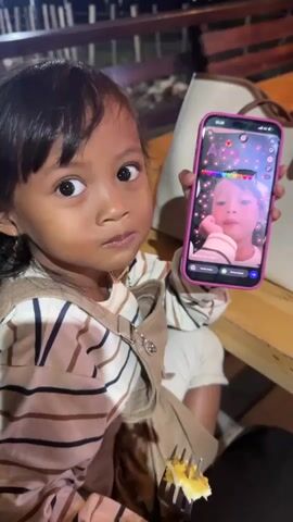 A young child with dark hair and brown eyes is sitting and holding a pink smartphone in their right hand. The phone's screen displays a selfie of the child with colorful, animated hearts and the word "Arka" written in rainbow colors. The child is wearing a striped shirt and brown overalls. The child is looking at the camera and appears to be speaking. In the foreground, a fork with a piece of cake is visible. The background suggests an outdoor setting at night with blurred lights.