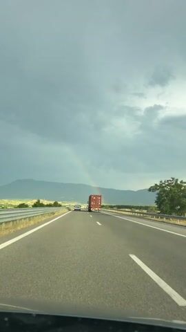 The video shows a view from inside a vehicle driving on a highway. The road ahead is visible, with a truck and a smaller vehicle in the distance. A rainbow arcs across the sky. The sky is overcast with dark clouds, and mountains are visible on the horizon. The environment appears to be rural.