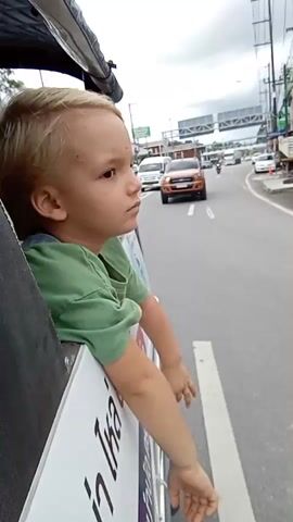 A young boy with blond hair and a green t-shirt is sitting in the back of a vehicle, possibly a tuk-tuk. He is looking out of the side of the vehicle at the road and other traffic. The boy's arms are resting on the side of the vehicle, and his hands are hanging down. The vehicle appears to be moving, as indicated by the blur of the road and the movement of other vehicles in the background. The environment seems to be an urban or semi-urban area with buildings and power lines visible. The boy's facial expression is thoughtful or perhaps a little bored. There is text visible on the side of the vehicle, but it is not clearly legible.
