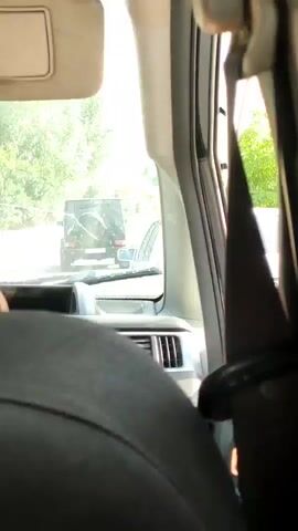 The video is shot from inside a vehicle, looking forward through the windshield. The perspective is from the back seat, focusing on the headrest and part of the passenger seat. Ahead, another vehicle, a dark-colored SUV with a spare tire mounted on the rear, is visible on a road. Trees and foliage line the sides of the road, indicating an outdoor, possibly rural or suburban setting. The lighting suggests it is daytime. The seatbelt of the vehicle is also visible on the right side of the frame.