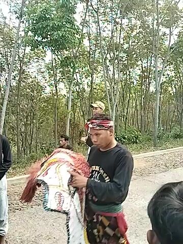 The video shows an outdoor gathering, possibly a street performance or a cultural event. In the foreground, a person's head is visible, looking down. The camera then pans to show a line of people sitting or standing along a dirt path, with trees in the background. Further down the path, a group of people are gathered around what appears to be a sound system and a decorated stage or truck. One person in the center is wearing a distinctive costume with colorful patterns and is standing near the sound equipment. The overall atmosphere suggests a lively event with spectators and performers.