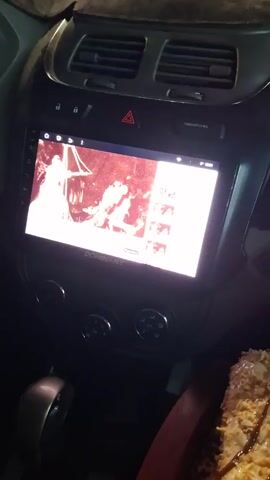 The video shows the dashboard of a car. A digital display shows what looks like a black and white video, possibly of musicians. There are three knobs beneath the screen and the hazard light button above. To the right of the gear shift, a dessert is visible. It appears to be a layered cake or pastry with a light-colored topping drizzled with chocolate sauce. The car's interior is dark and the lighting appears to be artificial. The camera moves slightly during the video.