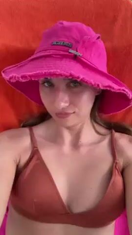 A young woman in a brown bikini top and a bright pink hat, presumably outdoors, is lying back in a beach chair, possibly taking a selfie. The woman is facing directly into the camera with a relaxed expression. The chair has an orange towel on it. Behind the woman, there's a glimpse of green grass and some plants, indicating an outdoor setting. The woman is not actively doing anything besides looking at the camera.