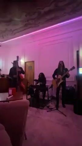 A band of three people performs in a softly-lit room. A man on the left is standing and playing a large stand-up bass. At the center, a figure wearing all black is playing a small drum kit. On the right, a man in black pants and shirt with long dark hair is playing an acoustic guitar and singing into a microphone. The room is lit with pinkish-purple light, and a neon strip light is mounted along the ceiling. A pink chair is visible in the lower left. A closed door is partially visible near the bass player. The band is arranged along the back wall and is playing with some energy.