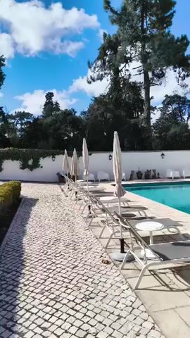 A bright, sunny day with scattered clouds is captured in this vertical video of a swimming pool area. The camera pans slowly across the scene. To the left, a cobblestone pathway is visible, lined with lounge chairs and closed umbrellas. The lounge chairs are made of white metal frames with grey fabric seats. White, minimalist chairs are also scattered around the area. In the center, a rectangular swimming pool with clear turquoise water is present. The pool is surrounded by a paved patio made of light-colored stone tiles. A white building with a red-tiled roof is situated behind the pool, featuring several windows and two green doors. Outdoor furniture, including white sofas and tables, is arranged in front of the building. Tall trees with green foliage stand behind the building, and more trees and foliage are visible along the white wall bordering the property on the left. The overall impression is one of a serene and well-maintained outdoor leisure space.