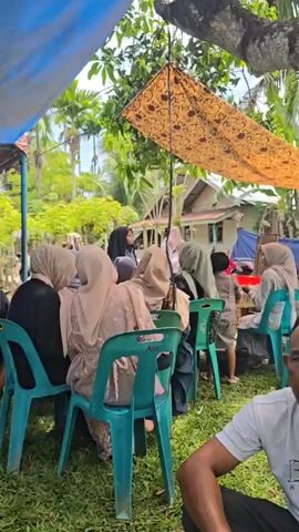 The video shows an outdoor gathering under a blue and brown tarp. Several people, mostly women wearing headscarves, are seated on teal plastic chairs arranged on a grassy area. Some people are eating at tables. In the background, there is a house and trees. The scene appears to be a social event or a celebration.