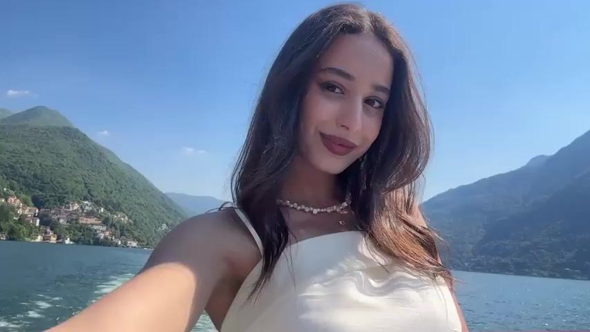 A young woman is taking a selfie on a boat. She has long brown hair, brown eyes, and is wearing a white dress and a pearl necklace. She is smiling at the camera. Behind her, there is a lake with mountains in the background. There is also a small town on the shore of the lake. The sun is shining brightly, and the sky is clear. The boat is moving, leaving a wake behind it.