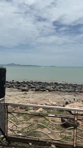 The video opens facing a body of water with a rocky shore in the foreground. A metal railing with a decorative design is visible in the lower portion of the frame. Beyond the shore, the sea stretches out, appearing calm with small waves. In the distance, landmasses or islands can be seen with a hazy sky overhead. A speed boat is visible in the middle ground, moving across the water and creating a wake. Two birds are also seen flying over the water. The camera pans slightly to follow the movement of the boat. The overall environment appears to be a coastal area on a slightly overcast day.