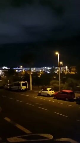 A nighttime view from an elevated position overlooking a street and a cityscape. The street in the foreground is paved, with white lane markings visible. Several vehicles are parked on the side of the street, including a white van, a white car, and a red car. A streetlamp illuminates the scene. In the mid-ground, there are trees and other vegetation. The background shows a city illuminated by numerous lights, spreading across hills under a dark, cloudy sky. The camera is static.