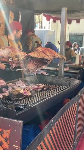 Here is a detailed description of the video:

The video shows a man cooking large cuts of meat at an outdoor market stall. The man wears glasses, a red cap, and a black apron, and he is working in front of a large grill. On the grill, there are numerous pieces of cooked meat. Above the grill, there are two large cuts of meat, each appearing to be half of an animal carcass, suspended from a metal bar. The meat is visibly charred and cooked. Behind the man, other individuals are working at the stall, and customers can be seen in the background. The stall is decorated in a rustic style, with dark wood accents. Menus with pricing are posted on the front of the stall. The general environment suggests a lively street food market or festival.