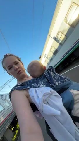 A woman in the foreground is filming a video on her phone, holding a baby in a carrier. Behind her, a train is stopped at a platform. To her right, two young girls are standing near a bench, and another woman is standing further down the platform, looking at her phone. The environment appears to be a train station platform.