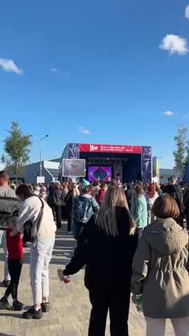 Here is a concise description of the video:

The video shows a crowd of people gathered in front of a stage. The stage is decorated with red, white, and blue colors. A large screen on the stage displays what appears to be live performance footage. The crowd is diverse, with people of different ages and wearing various styles of clothing. The sky is blue with a few clouds, indicating it is likely daytime. The setting seems to be an outdoor event or festival.