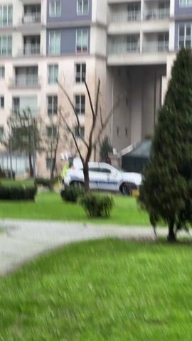 The scene shows a traffic police vehicle parked on a grassy area next to a walkway. The car is white with blue stripes and the words "TRAFIK POLISI" are visible on the side. A person wearing a yellow vest can be seen near the open back of the vehicle. In the background, there is a large building with multiple windows and balconies, and a black tent-like structure. A few small trees and bushes are scattered around the lawn. The weather appears to be overcast or rainy.