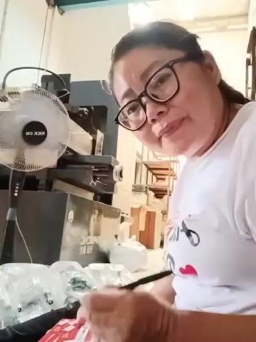 A woman wearing glasses and a white t-shirt with red hearts is writing with a black pen. She is sitting in front of a large, dark machine and a black oscillating fan. There are plastic-wrapped items and white bags on the floor in front of her. In the background, there are shelves with boxes. The scene appears to be indoors, possibly in a workshop or storage area.